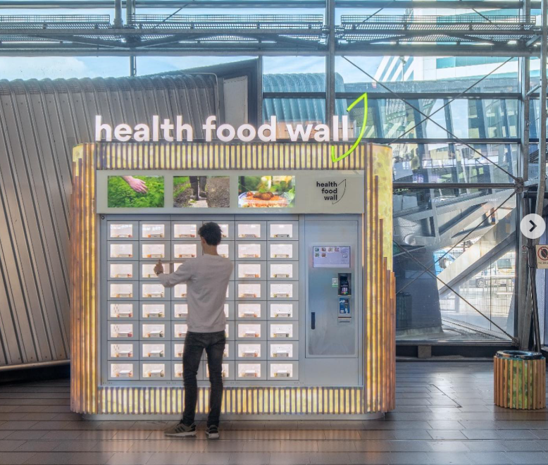 Person selecting a meal from a large Health Food Wall automated kiosk with multiple refrigerated cubbies.