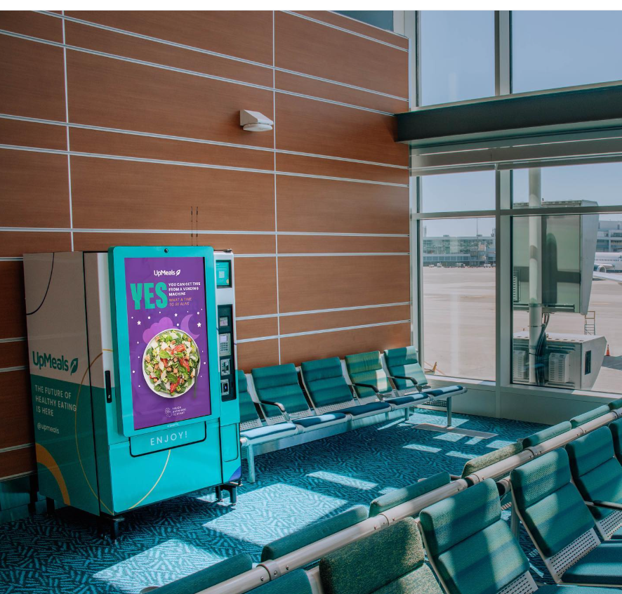 Teal UpMeals SmartVending machine located in a Vancouver Airport departure lounge, displaying a fresh salad on its digital screen as a healthy airport food vending machine option.